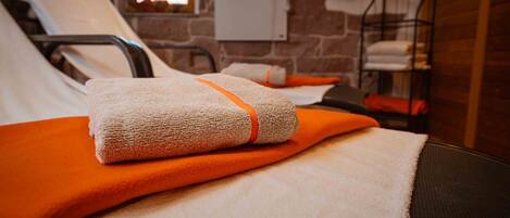 Ein Bett mit einem weißen Handtuch und einer orangefarbenen Decke, ein Metallregal mit Handtüchern und eine Backsteinwand.