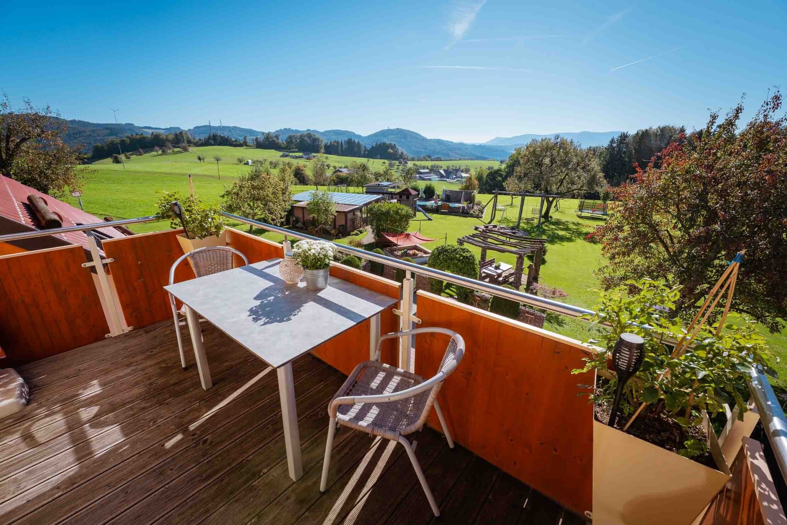 Un balcon avec une table et des chaises, donnant sur un paysage verdoyant parsemé de maisons et de collines.
