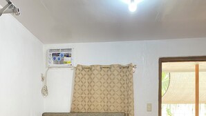 Ein Einzelbett mit Kopfteil, weißen Kissen, einem aufgerollten Handtuch und einer karierten Decke. An der Wand ist eine Klimaanlage angebracht, und ein Vorhang mit geometrischem Muster ist vorhanden.