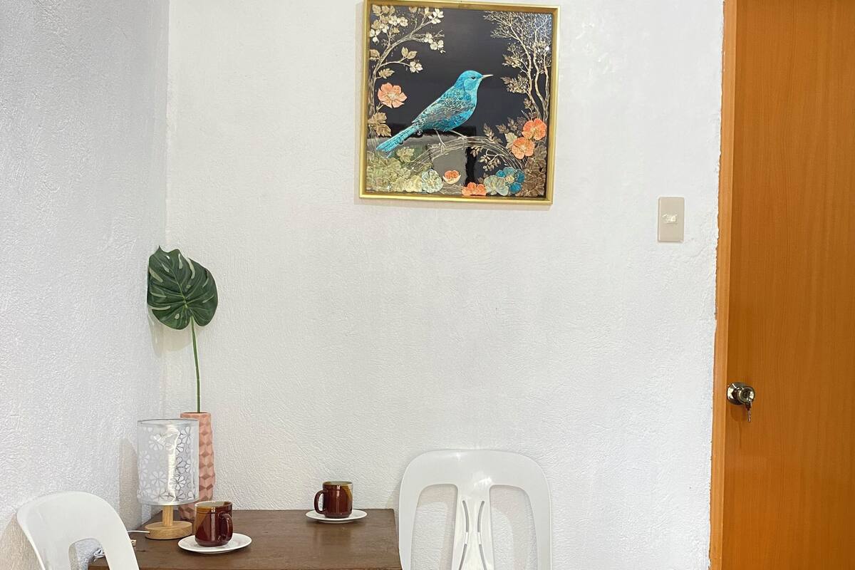 Ein kleiner Essbereich mit einem Holztisch, zwei weißen Plastikstühlen, einem Topfpflanzen und einem Bilderrahmen mit einem Vogelmotiv an der Wand.