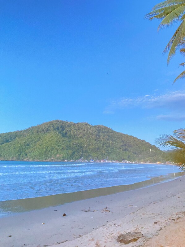 A beach with clear blue water, a sandy shore, and a hill with dense vegetation in the background. - Jasmine's Inn at Bucana Beach (El Nido)