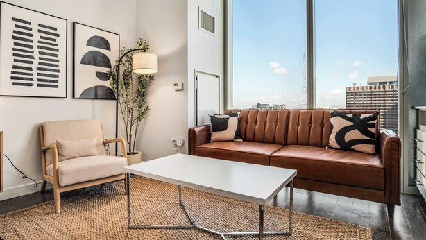 Een moderne woonkamer met een bank, fauteuil en salontafel. Er hangen kunstwerken in lijsten aan de muur, een vloerlamp en een groot raam met uitzicht op de stad.
