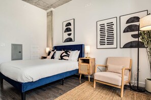 Un dormitorio moderno con una cama grande, cabecera azul, mesita de noche de madera, una silla y una obra de arte en la pared.