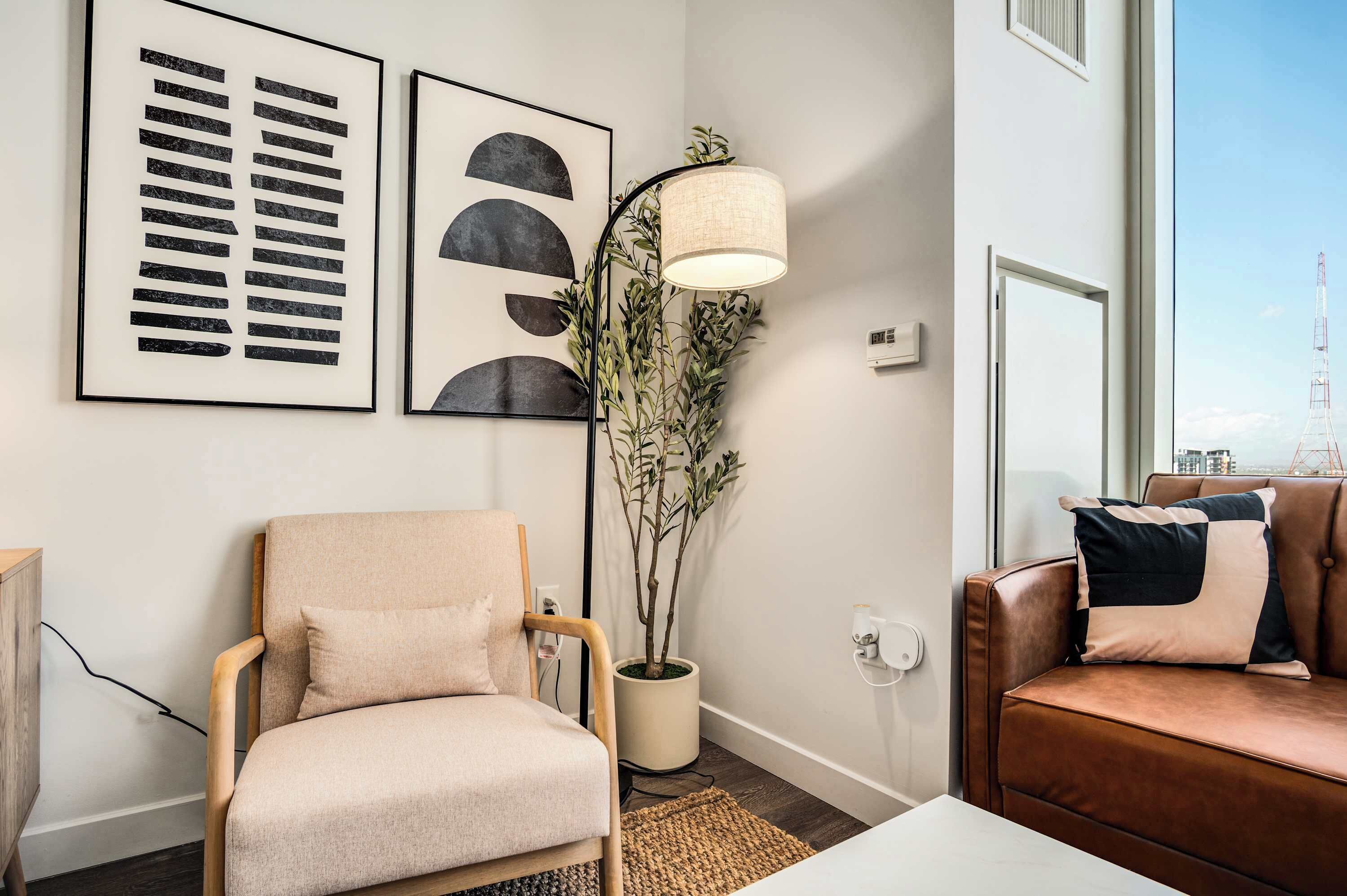 A living room with a beige armchair, a brown sofa, a floor lamp, and two framed abstract artworks on the wall.