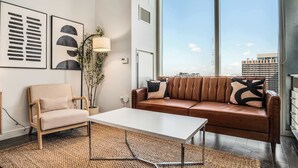 A modern living room with a sofa, armchair, and a coffee table. There are framed artworks on the wall, a floor lamp, and a large window with a city view. - Boulevard Stays Highrise STU Sky Pool, Parking, Gym 26 (Phoenix)