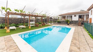 Ein rechteckiger Pool mit Steinbord, umgeben von einer Steinmauer und einer Pergola mit weißen Stühlen.