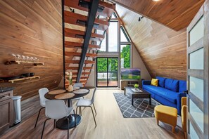 Un acogedor loft con interior de madera, una zona de comedor con una mesa y sillas redondas, un sofá azul y una escalera que sube a un nivel superior.