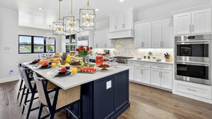 Una cocina moderna con armarios blancos, una isla central con frutas y flores, electrodomésticos de acero inoxidable e iluminación colgante.