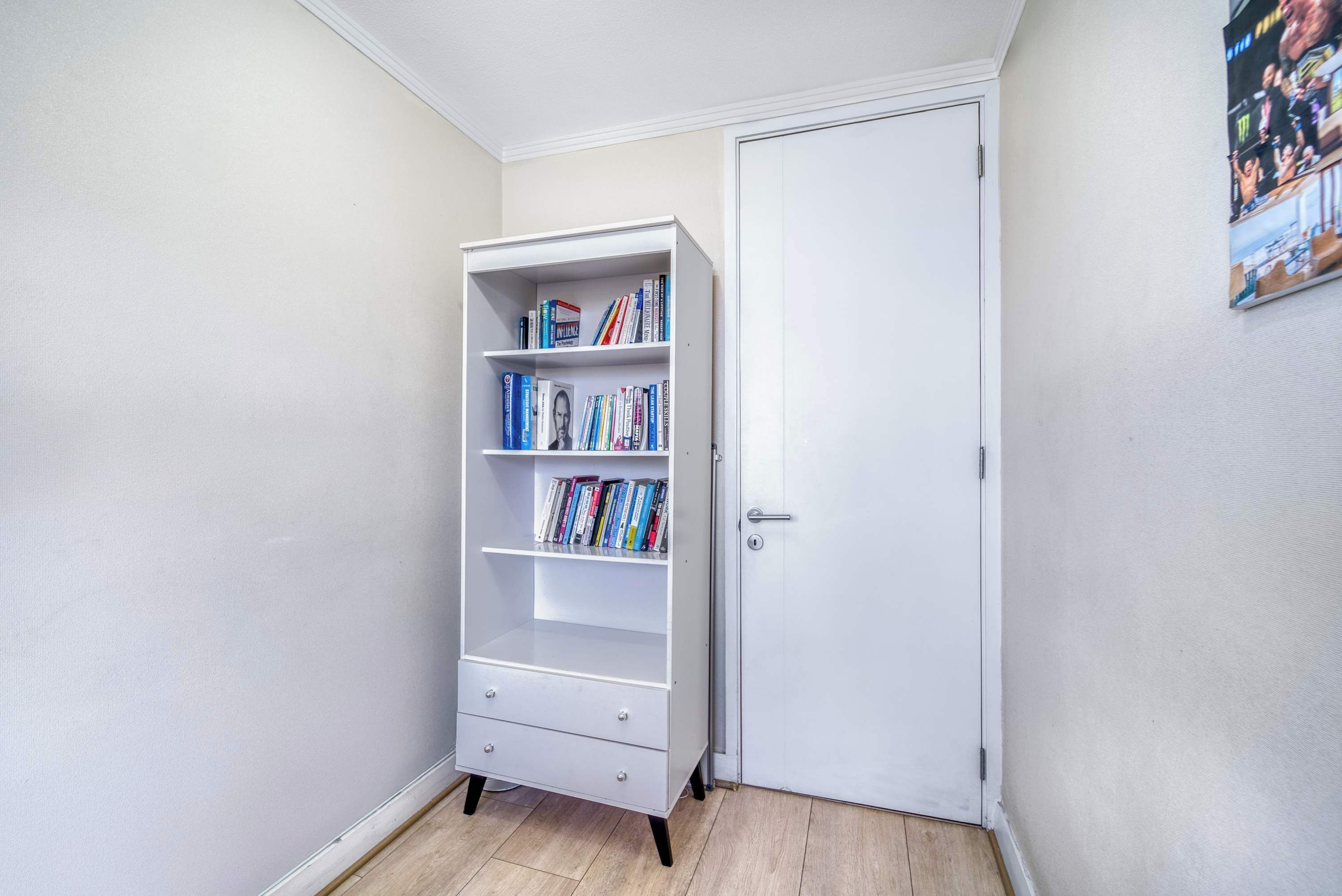 Quarto de canto com uma estante branca, uma porta e um cartaz na parede.