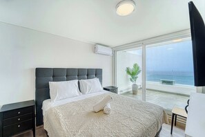 A bedroom with a large bed, a bedside table, a nightstand, a potted plant, and a view of the sea.