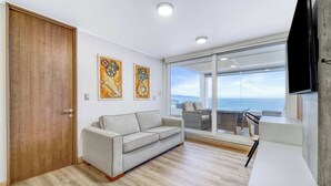 A modern living room with a sofa, wooden flooring, and a large window offering a view of the sea. - Infinite Coast 2BR with pool, gym & free parking (Viña del Mar)