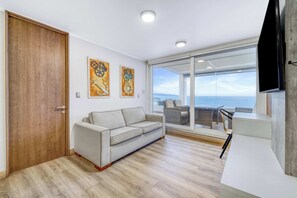 A modern living room with a sofa, wooden flooring, and a large window offering a view of the sea. - Infinite Coast 2BR with pool, gym & free parking (Viña del Mar)