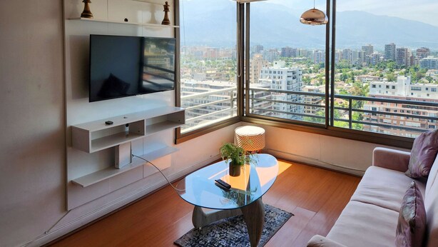 A modern living room with a TV, a glass coffee table, a sofa, and a large window offering a city view. - La Gloria Home - Comfort and City Views (Las Condes)