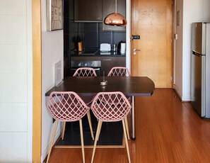 Une cuisine compacte comprenant une table à manger, quatre chaises roses, un réfrigérateur en acier inoxydable et un luminaire suspendu en cuivre.