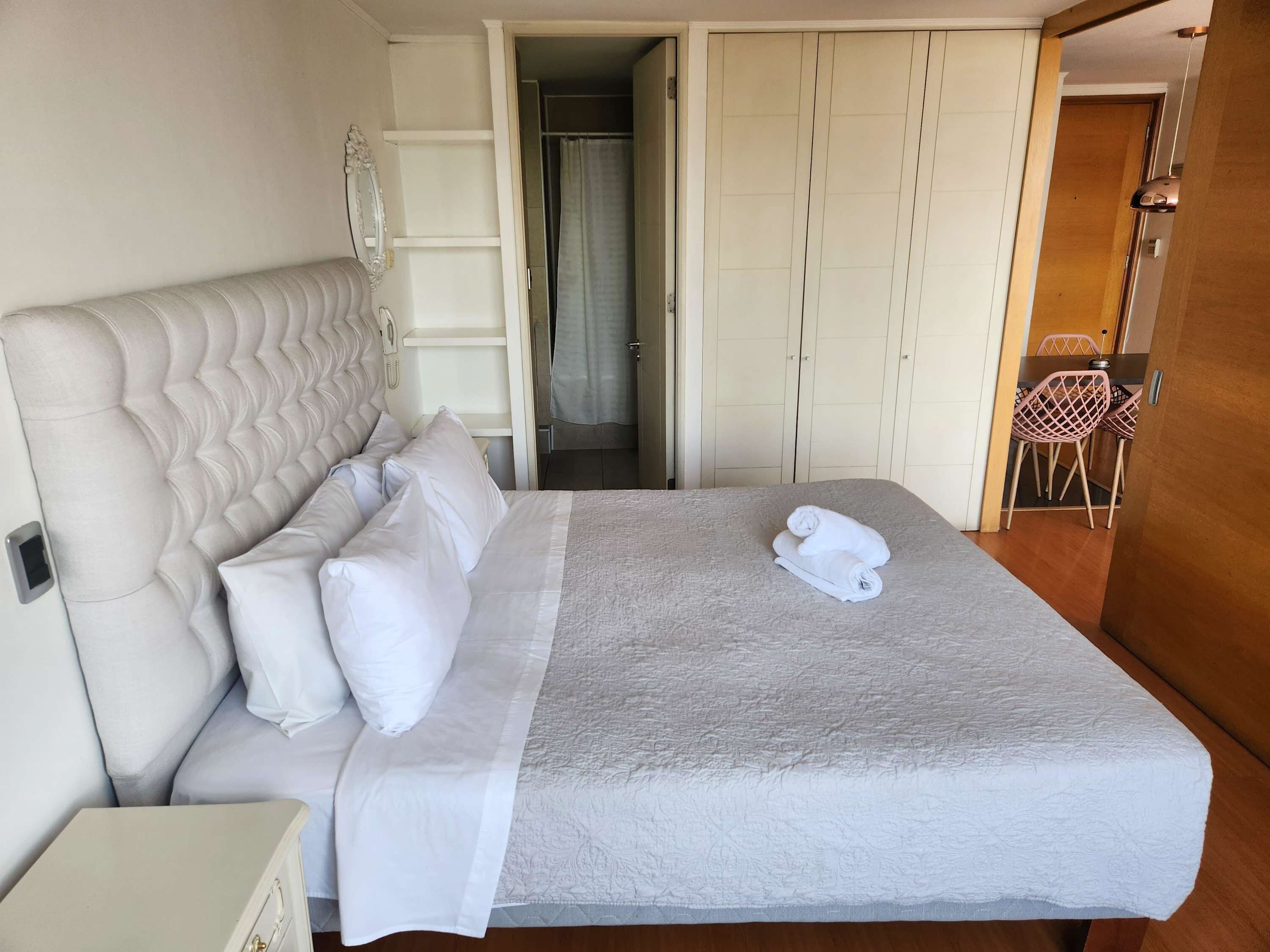 Una cama bien hecha con almohadas, una mesita de noche y una silla en una habitación con un armario y una puerta que da a otra habitación.