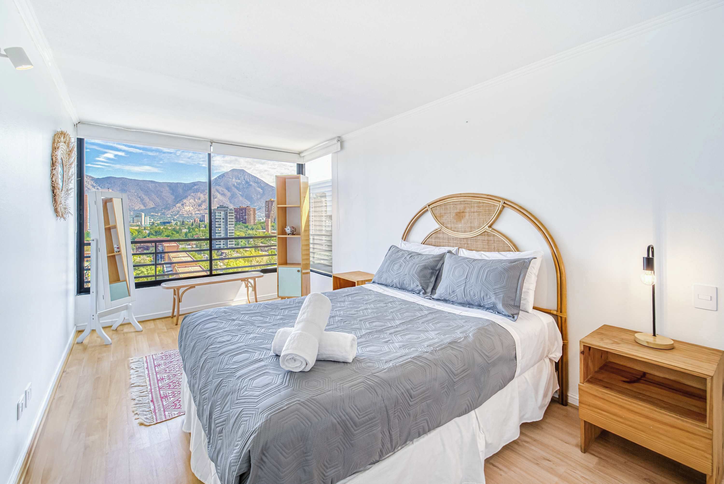 Ein Schlafzimmer mit einem großen Bett, einem Nachttisch, einer Lampe, einem Fenster mit Blick auf Gebäude und Berge sowie einem kleinen Tisch mit einem Stuhl.