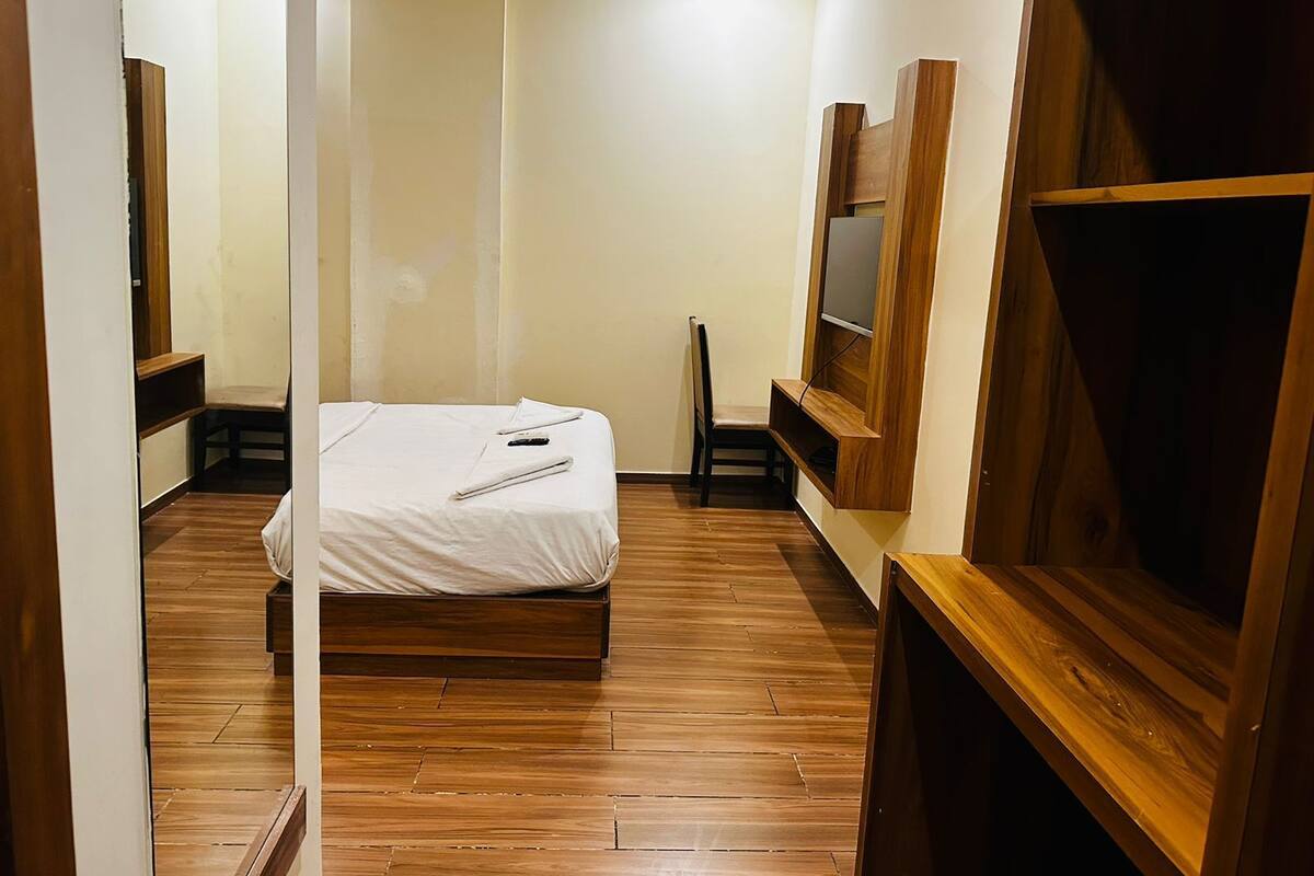 A bedroom with a wooden floor, a bed, a chair, a TV mounted on the wall, and a wooden cabinet.