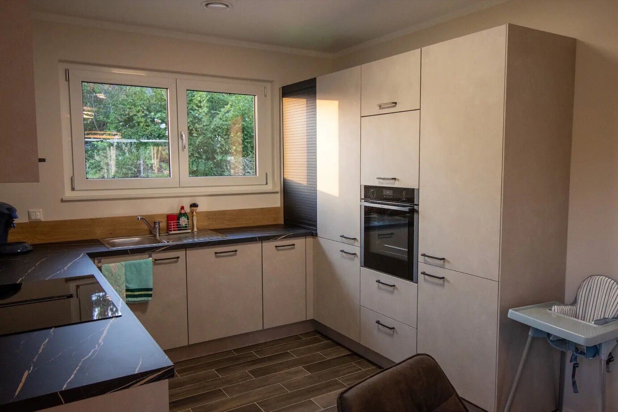 A modern kitchen with a built-in oven, a window with a view of greenery, and a child's high chair.