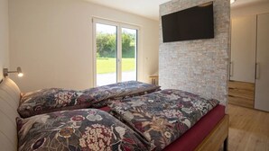 A bedroom with a bed, a TV mounted on a brick wall, and a sliding glass door leading to a garden.