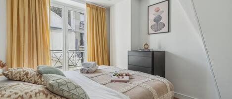 Ein Schlafzimmer mit Bett, Kissen, Nachttisch, einem gerahmten Bild und einem Fenster mit Vorhängen.