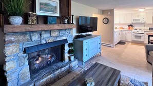 Una sala de estar con chimenea de piedra, un televisor de pantalla plana y una cocina al fondo.