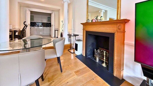 Een moderne woonkamer met een glazen eettafel, witte stoelen, een open haard en een grote spiegel.