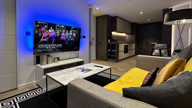 Ein modernes Wohnzimmer mit Flachbildfernseher, einer gelben Kissen auf dem Sofa, einem Couchtisch und einer Küchenzeile im Hintergrund.