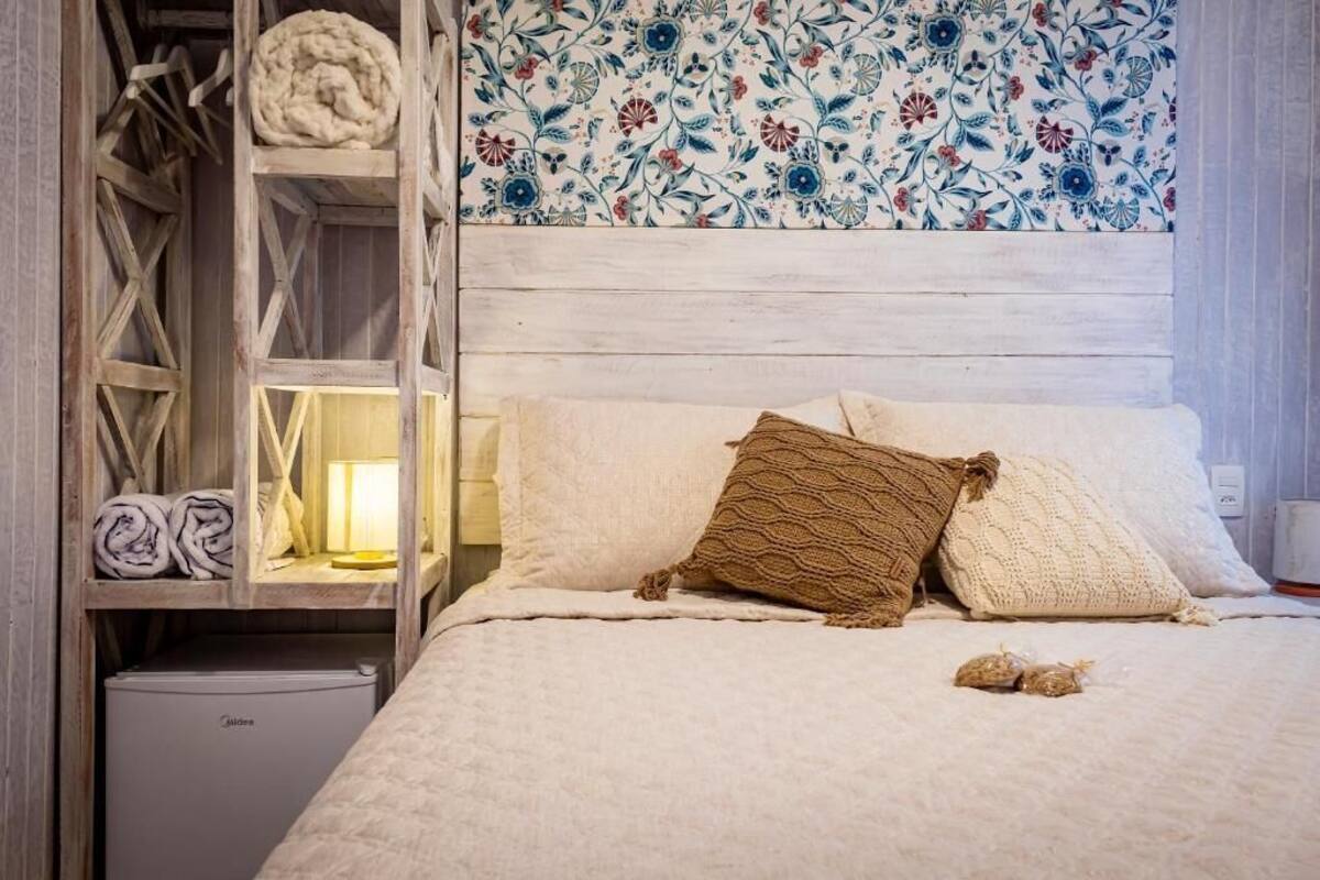A bedroom with a wooden headboard, a bed with white bedding and two decorative pillows, a small bedside table with a lamp, and a built-in wardrobe with a floral-patterned wallpaper.