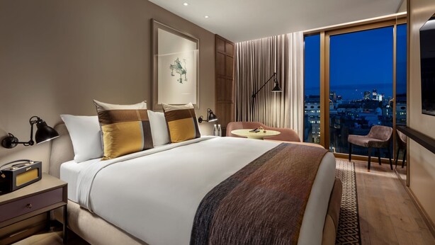 A modern hotel room with a large bed, a desk with a lamp, a chair, and a view of the city at night. - Uranium Continental Hotel (Kaduna)