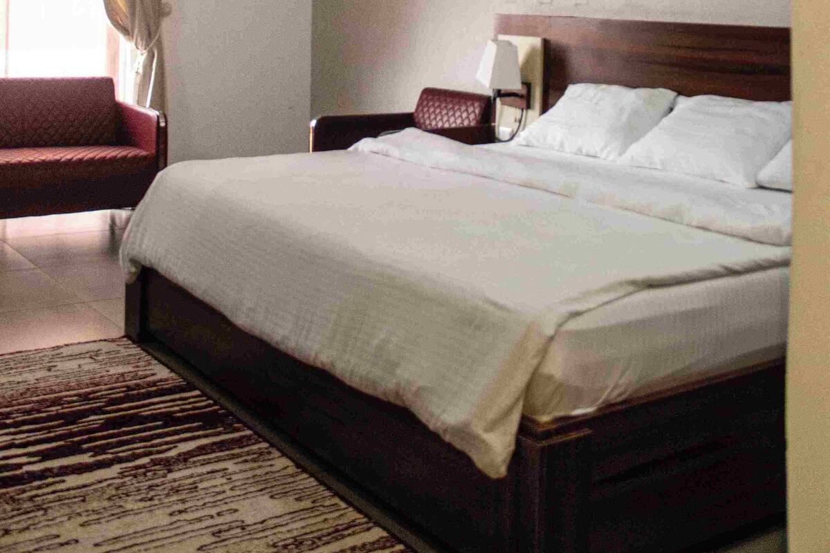 Une chambre d’hôtel avec un grand lit, une table de chevet avec une lampe, une chaise et une fenêtre avec des rideaux.