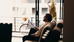 Una persona sentada en un sofá en una habitación con ventanas amplias, una bicicleta afuera y una planta en el alféizar.