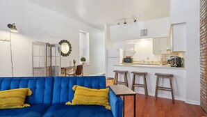 A modern living room with a blue sofa and yellow cushions, a small table, and a kitchen area with a bar stool. - Charming Soulard Retreat With Historic Character 1 Bedroom Apts (St. Louis)