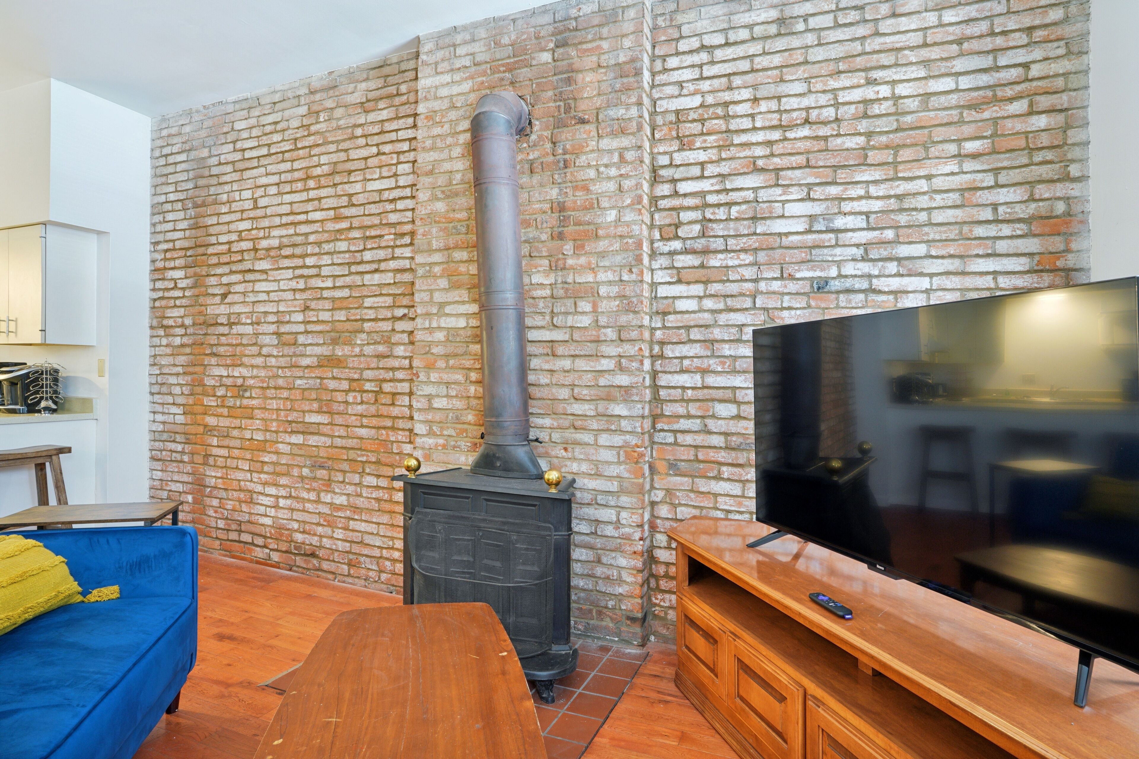 A living room with a brick wall, a black wood-burning stove, a blue sofa, a wooden table, and a TV on a wooden stand.