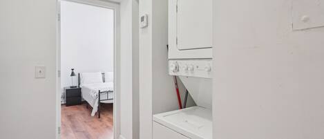 A laundry room with a washing machine and dryer, a bed in the adjacent room, and a door leading to another area.