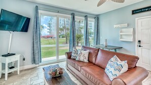 A living room with a brown leather sofa, a flat-screen TV, a coffee table with decorative items, and a ceiling fan. - Nantucket Cottage 06ab 2 Bedroom Condo (Destin)