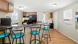 Une cuisine avec des meubles de rangement en bois, un réfrigérateur noir, un four à micro-ondes et une machine à café. On y trouve trois tabourets de bar avec des coussins turquoise.