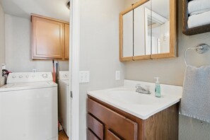 Une petite salle de bain équipée d’un lave-linge, d’un lavabo avec un distributeur de savon et d’un miroir au-dessus du lavabo.
