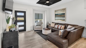 Un salon moderne avec un canapé en cuir marron, un fauteuil à motifs, un téléviseur à écran plat fixé au mur et un ventilateur de plafond.