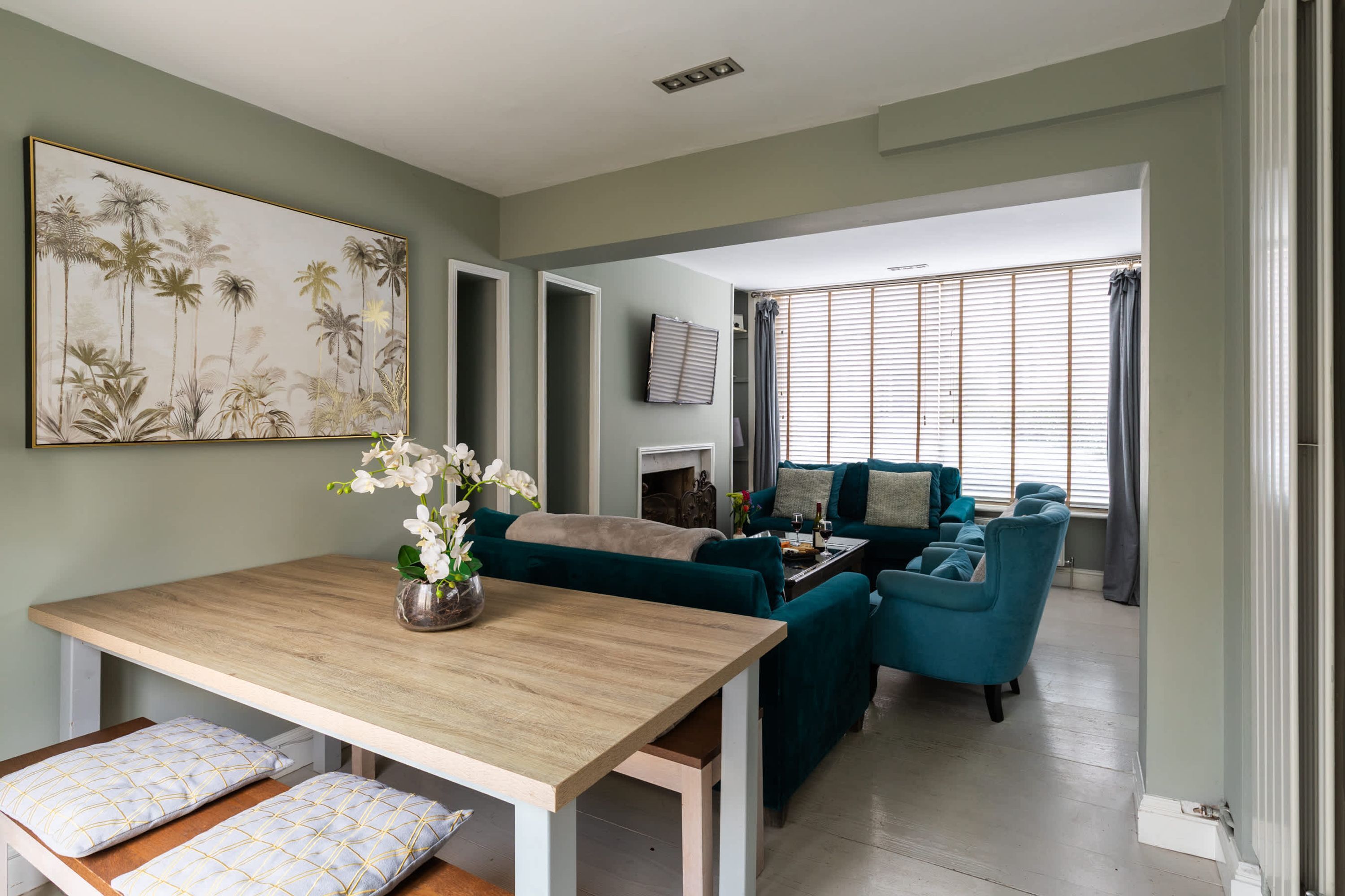A spacious living area with a wooden dining table, teal sofas, and a large wall-mounted painting.