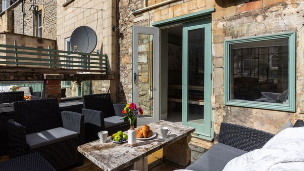 An outdoor seating area with wicker furniture, a stone table with a vase of flowers, and a view of a stone building with a door and window. - Stylish 4-bedroom Townhouse in the Heart of Bath (Bath)