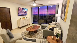 A modern living room with a large window, a flat-screen TV, a wooden coffee table, and a sofa. - Stunning Condo With Panoramic Ocean City Views (Cabo San Lucas)