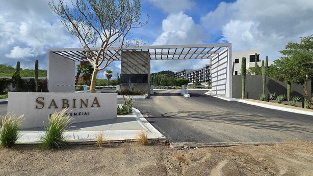 A sign for 'Sabina Financial' with a modern architectural entrance, a clear sky, and cacti in the foreground. - Stunning Condo With Panoramic Ocean City Views (Cabo San Lucas)