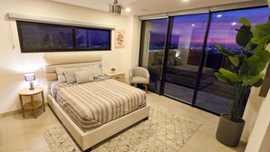A bedroom with a bed, bedside table, lamp, and a large window with a city view. - Stunning Condo With Panoramic Ocean City Views (Cabo San Lucas)