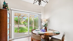 A dining area with a table set for two, a chandelier, and a view of a garden through a sliding glass door.