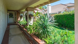 A covered walkway with a brick border, leading to a house with a tiled roof and a well-maintained garden with various plants and a large agave plant.