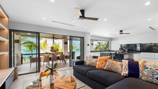 A modern living room with a glass coffee table, a dark sofa with patterned cushions, and a view of the ocean through large sliding doors. - Newly Renovated Notos Cove Villa Property (Nassau)
