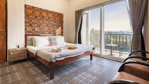 Ein Schlafzimmer mit einem großen Bett, einem Holz-Kopfteil, einem gemusterten Boden, einem Balkon mit Ausblick und einem kleinen Tisch mit Stuhl.