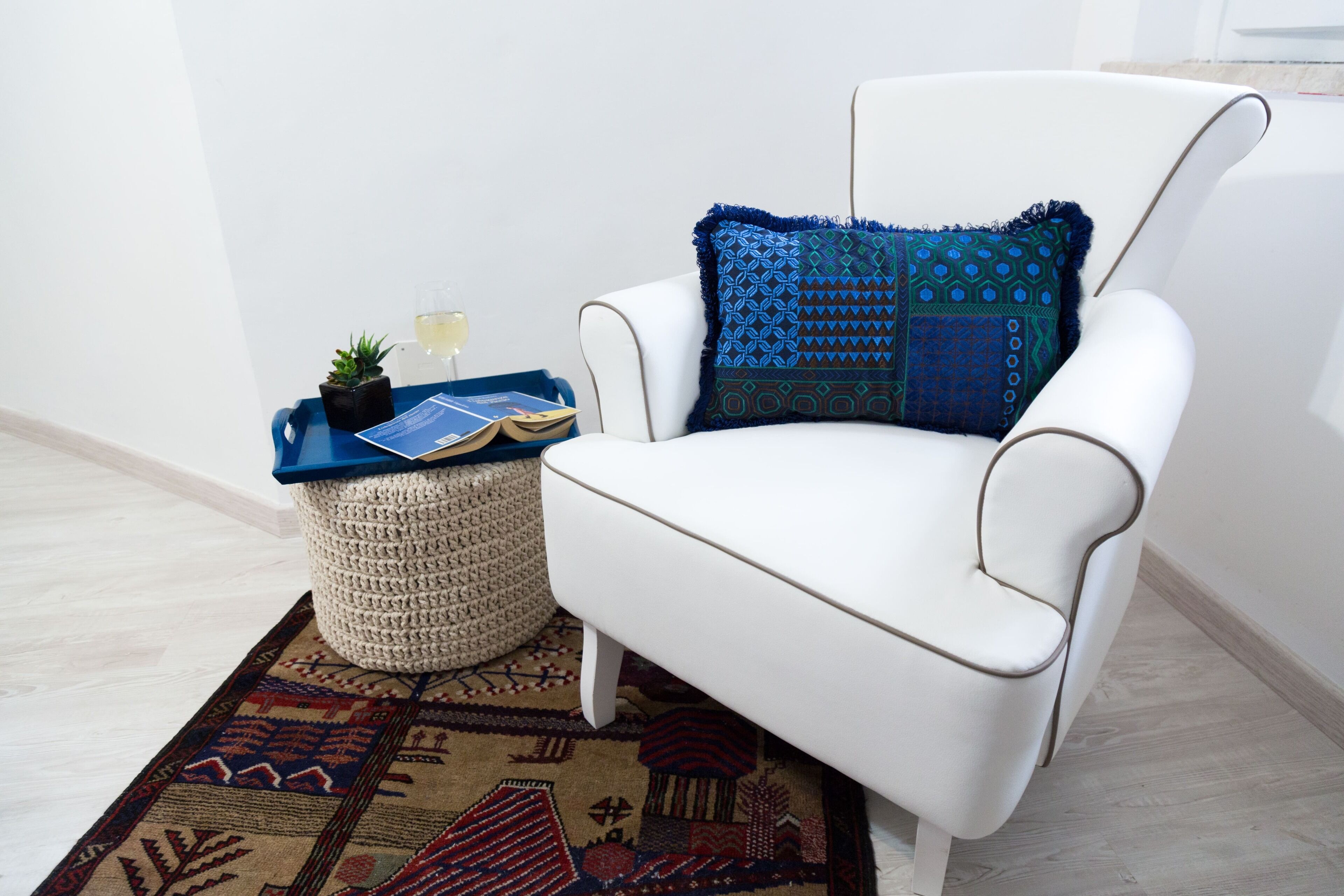 A white armchair with a patterned cushion, a small side table with a tray, a potted plant, and a woven ottoman.