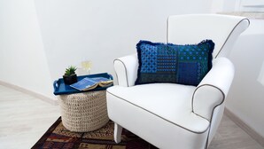 Un sillón blanco con un cojín estampado, una mesita auxiliar con una bandeja, una planta en maceta y un otoman tejido.