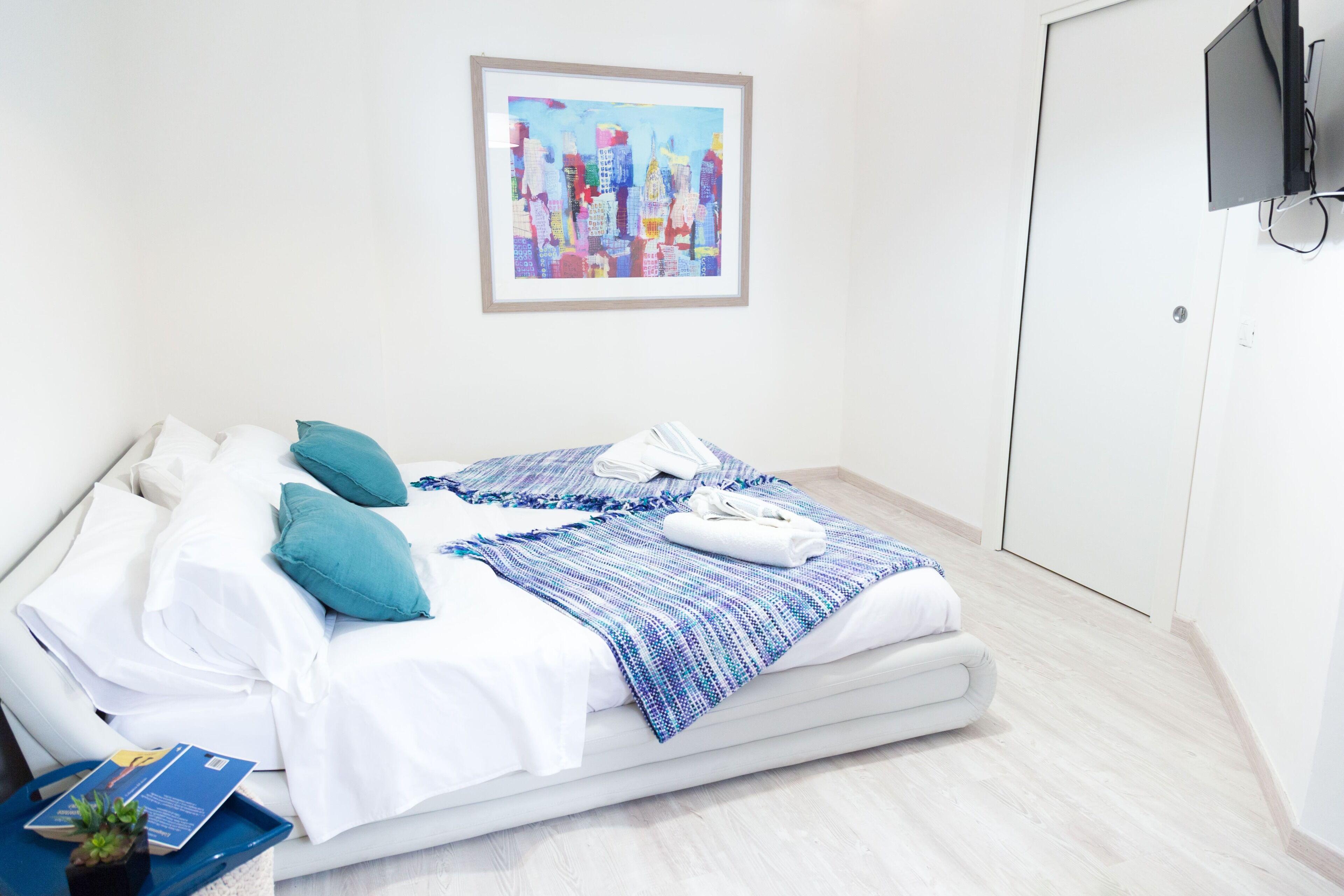 Un lit bien fait, avec des draps blancs, des couvertures à motifs bleus et blancs, et des coussins turquoise. Un tableau représentant un paysage urbain, encadré, est accroché au mur au-dessus du lit.